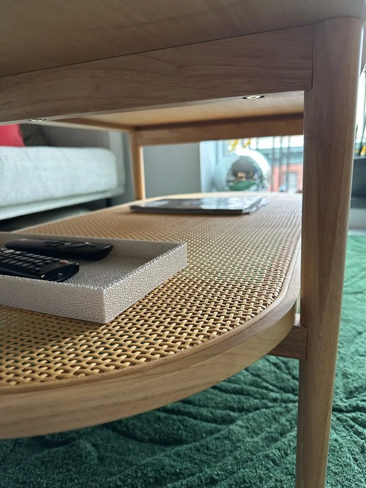 Closeup of the rattan lower shelf of an oval two-tier coffee table within a living room. 