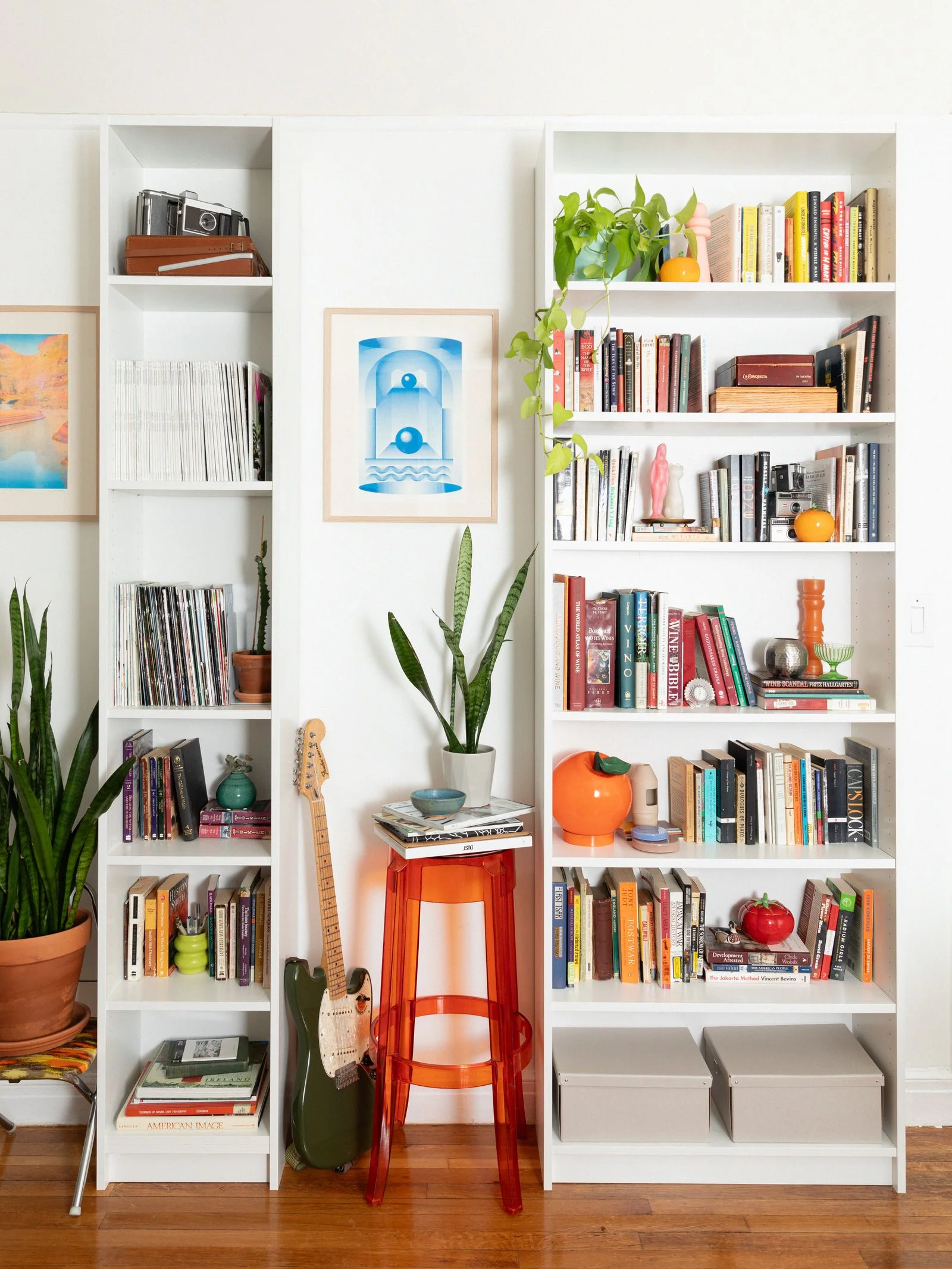 Two white IKEA bookshelves in a living room one narrow and one wide styled with various books and knick knacks.