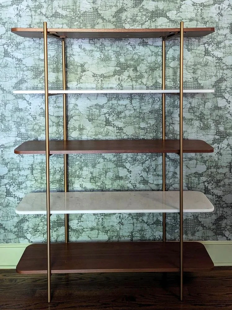 5-tier bookshelf with alternating wood and marble shelves and a gold metal frame in front a green wallpapered wall.