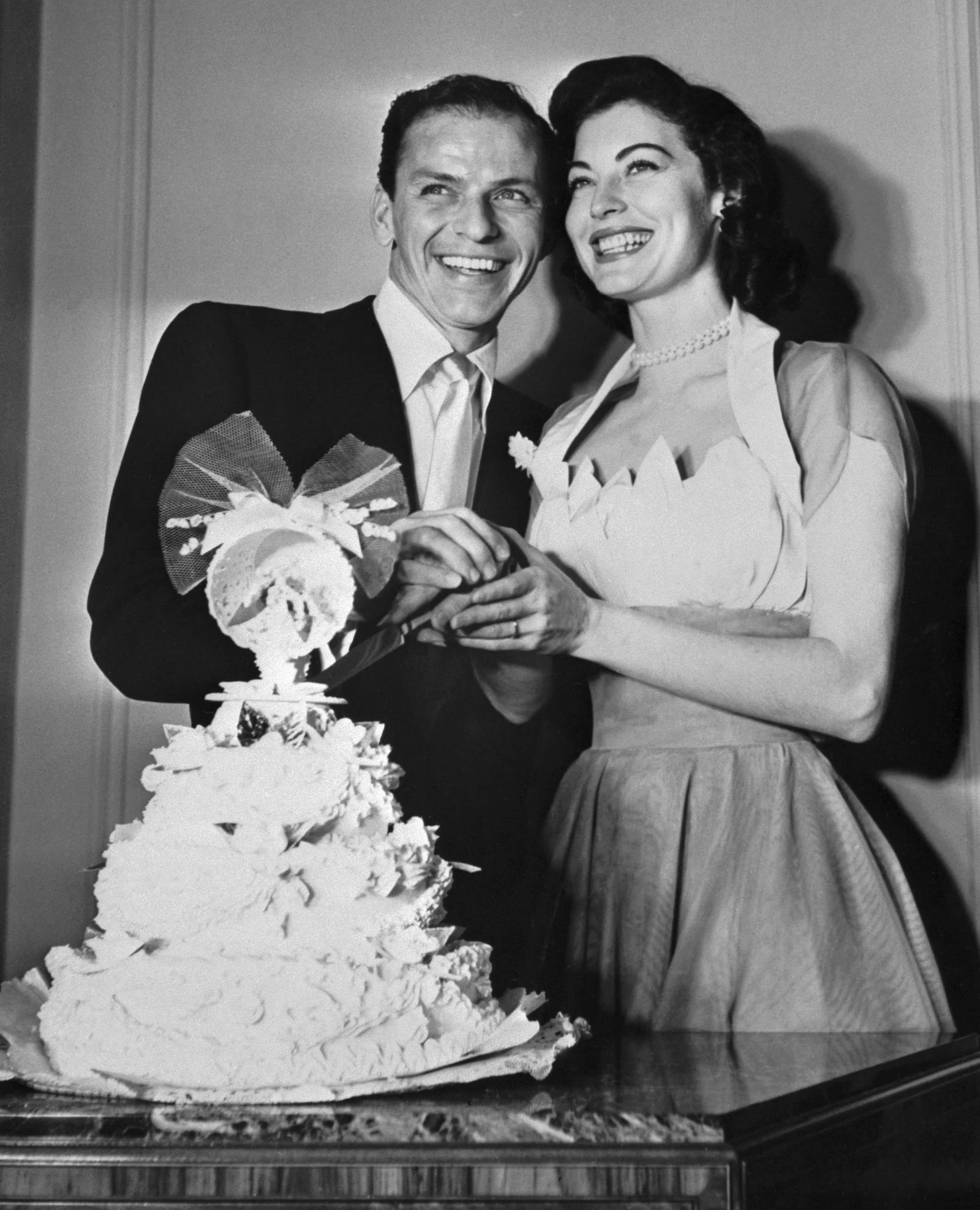 blackandwhite photo of Ava Gardner right posed with Frank Sinatra both smiling behind their tiered wedding cake