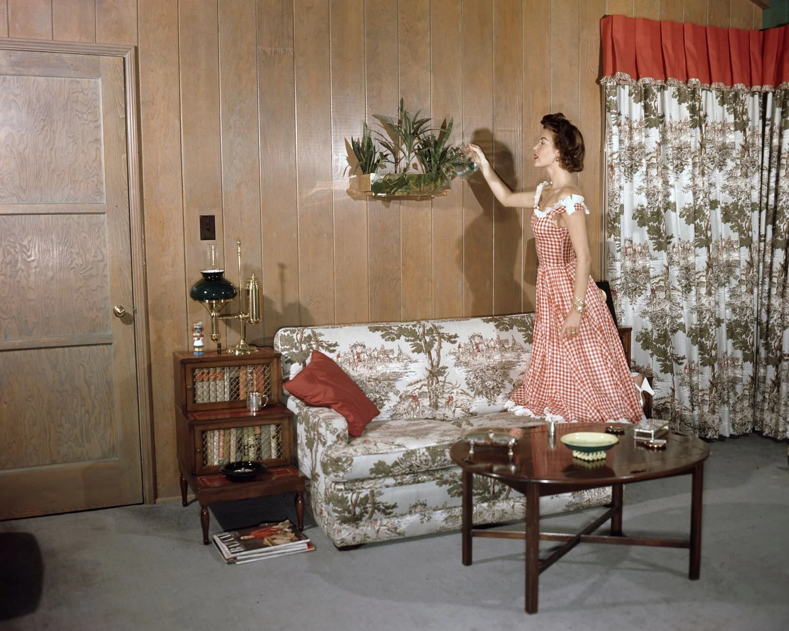 Ava Gardner in red and white gingham dress tending to plant mounted on shelf on wood paneled wall gray carpet matching...