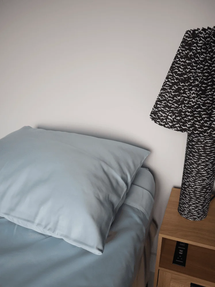 Bed made with blue sheets next to a nightstand with a black and white lamp on it.