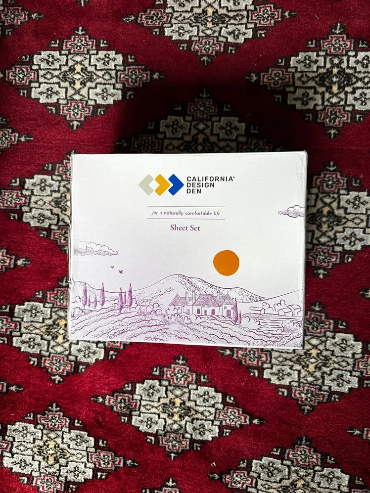 Opened box of California Design Den sheets on a red patterned rug.